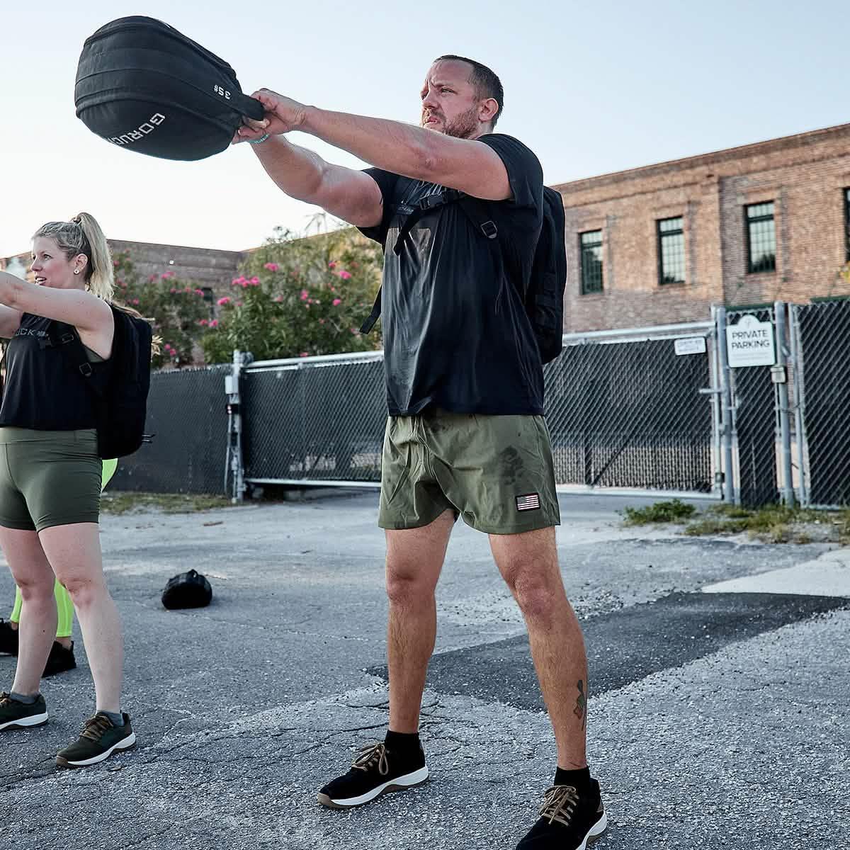 Man wearing GORUCK gear and USA ranger green training shorts exercising outdoors with kettlebell near brick building