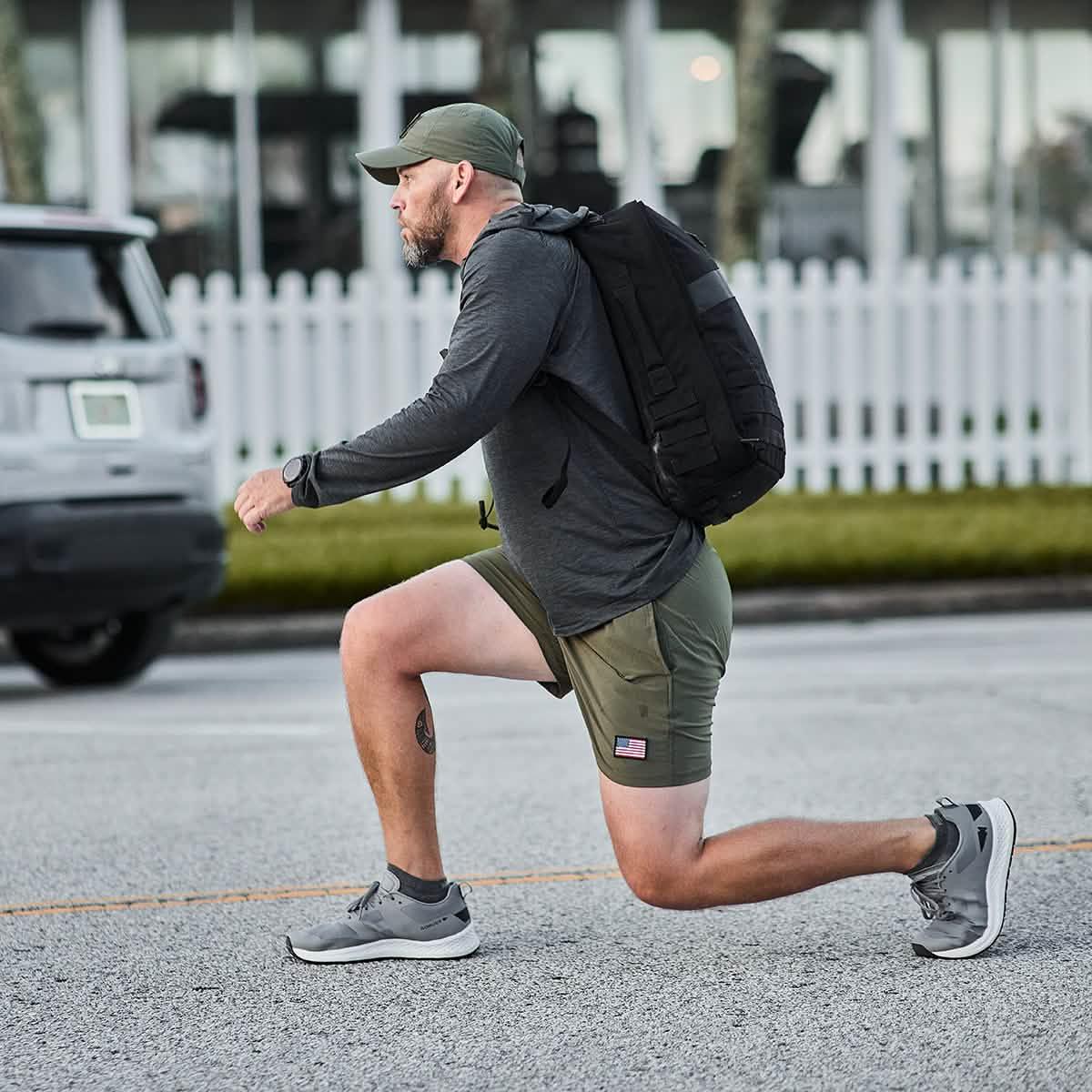 Man in ranger green USA training shorts lunging outdoors wearing black backpack and gray hoodie