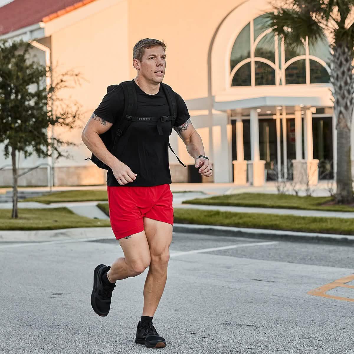 Man running outdoors wearing black GORUCK shirt, red athletic shorts, and black sneakers with backpack