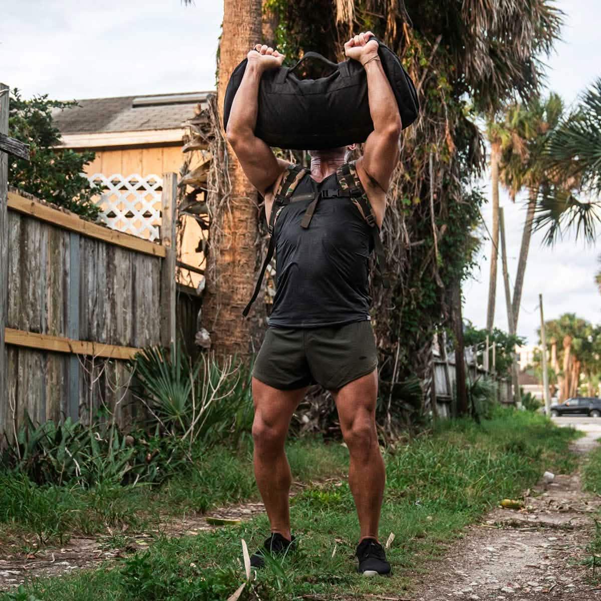 Muscular man in black tank top and shorts lifting weighted rucksack outdoors near wooden fence and palm trees