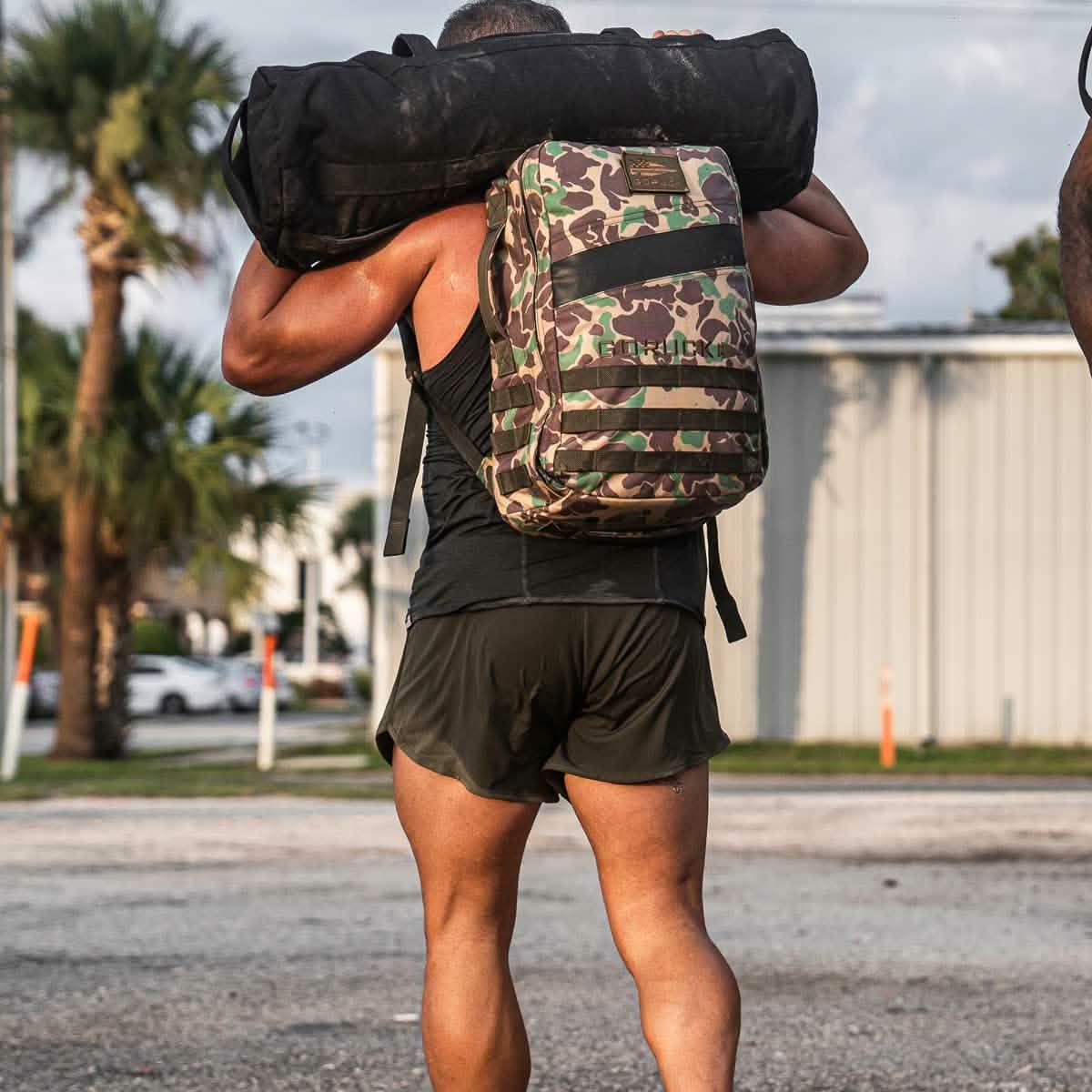 Man wearing camo GORUCK backpack and black workout shorts carrying heavy sandbag outdoors
