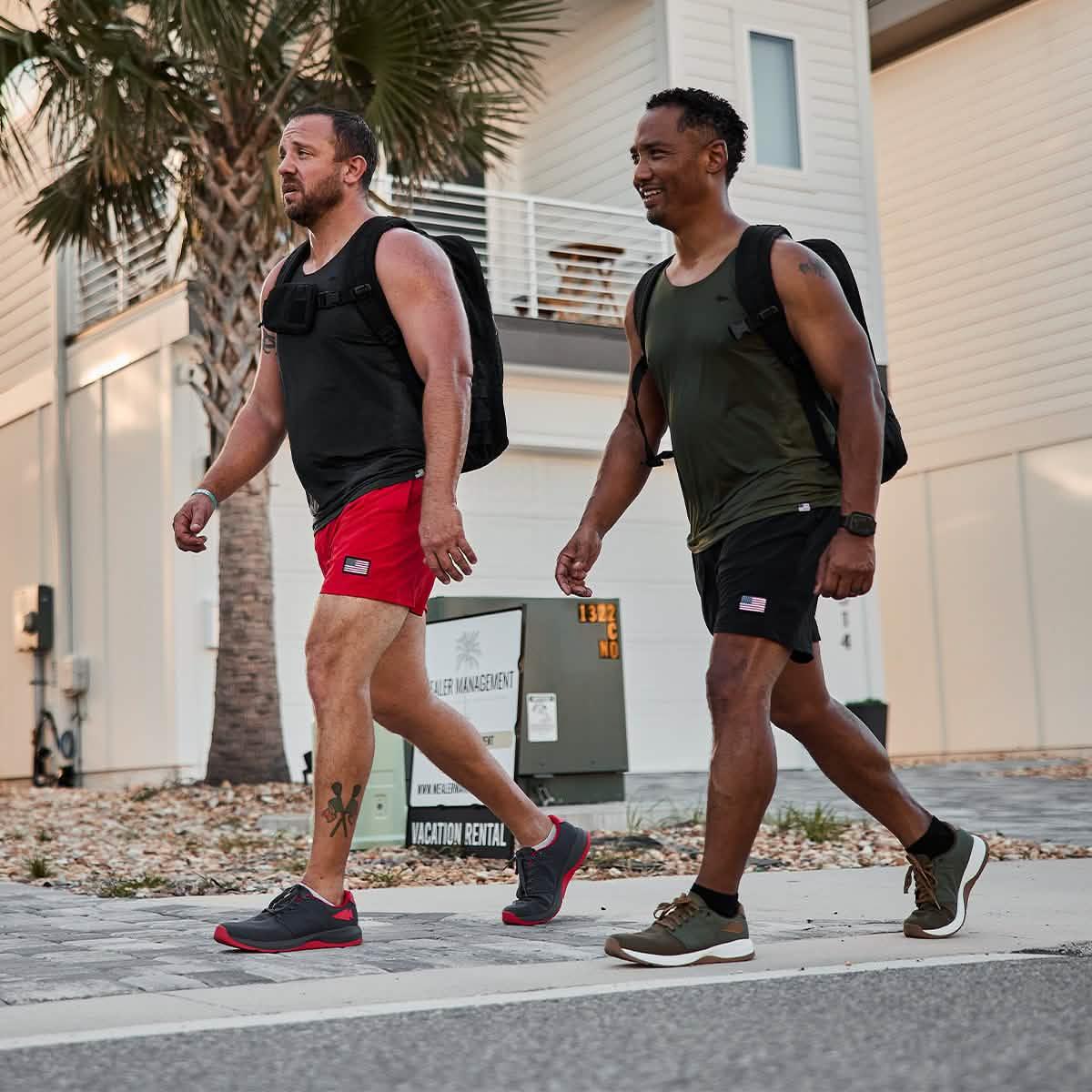 Two men walking outdoors wearing GORUCK black and green tank tops with American flag shorts and backpacks