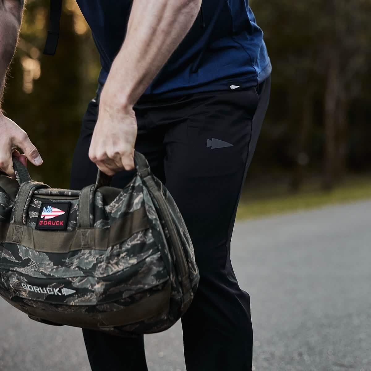 Close-up of a man wearing black GORUCK joggers, holding a camouflage GORUCK weighted bag outdoors