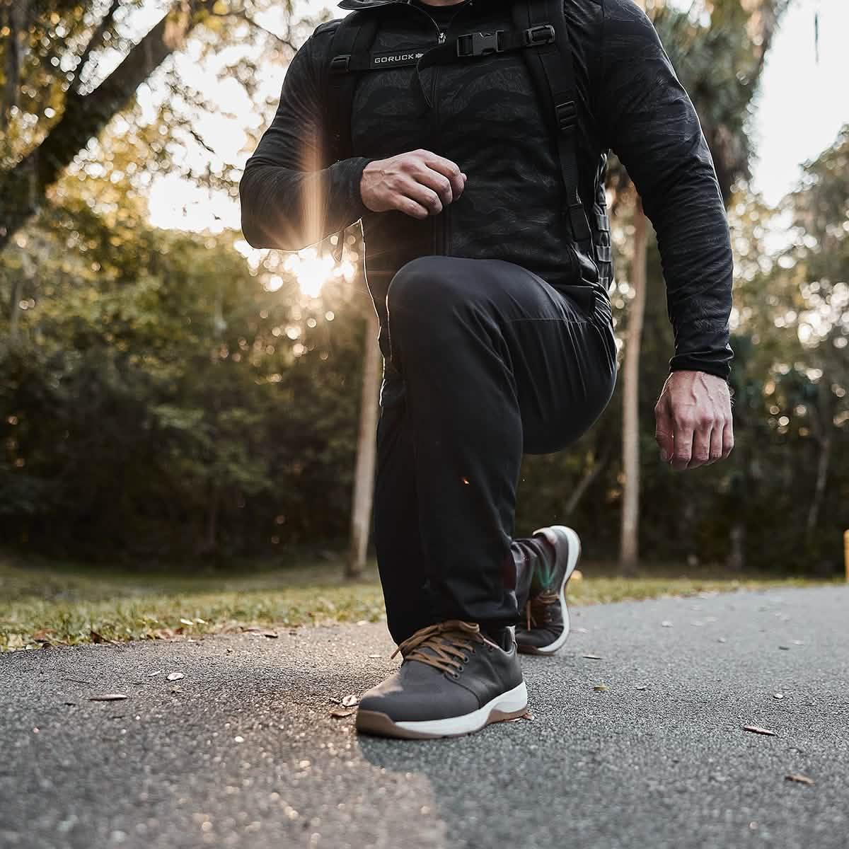 Person wearing black GORUCK joggers and sneakers lunging outdoors on a sunlit forest path