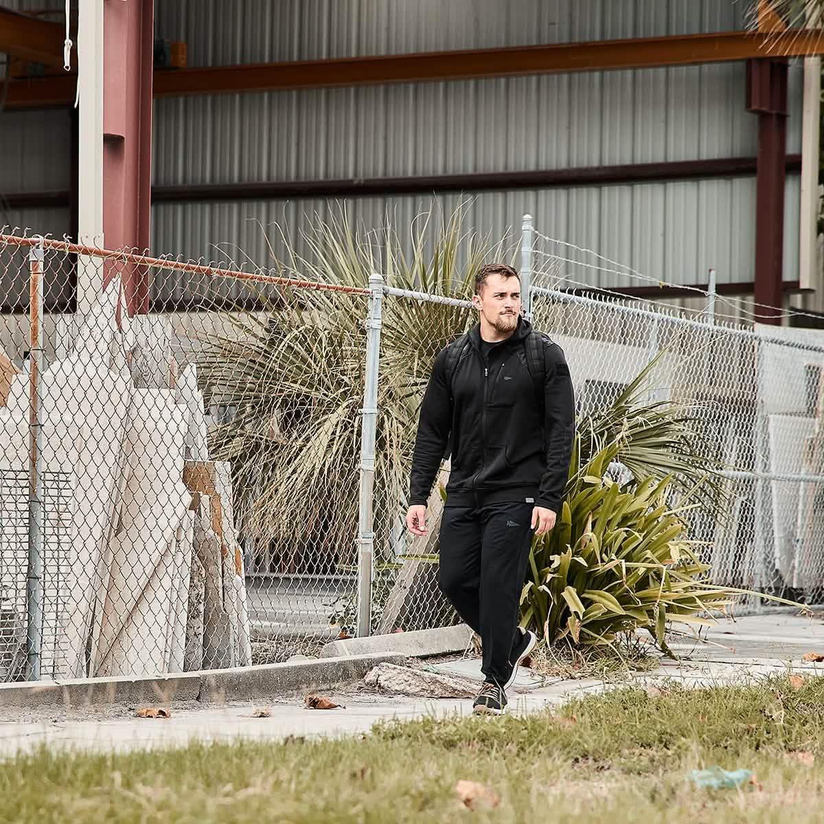 Man wearing GORUCK black joggers and hoodie walking outdoors by chain-link fence and industrial building