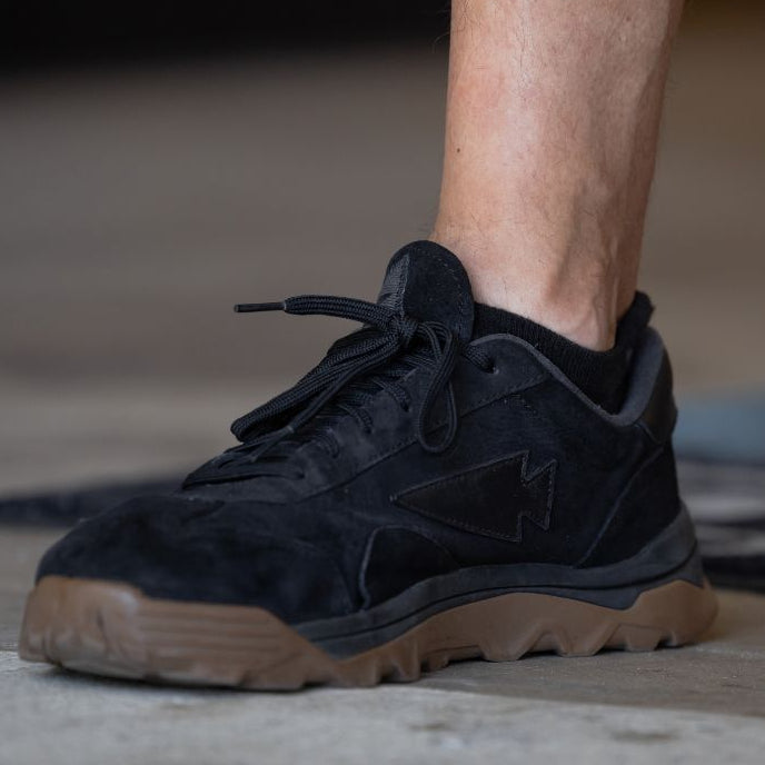 A person stands on a floor with only one black sneaker with brown sole and lower leg visible, wearing Merino Challenge Socks - Ankle (No-Show) made in the USA.