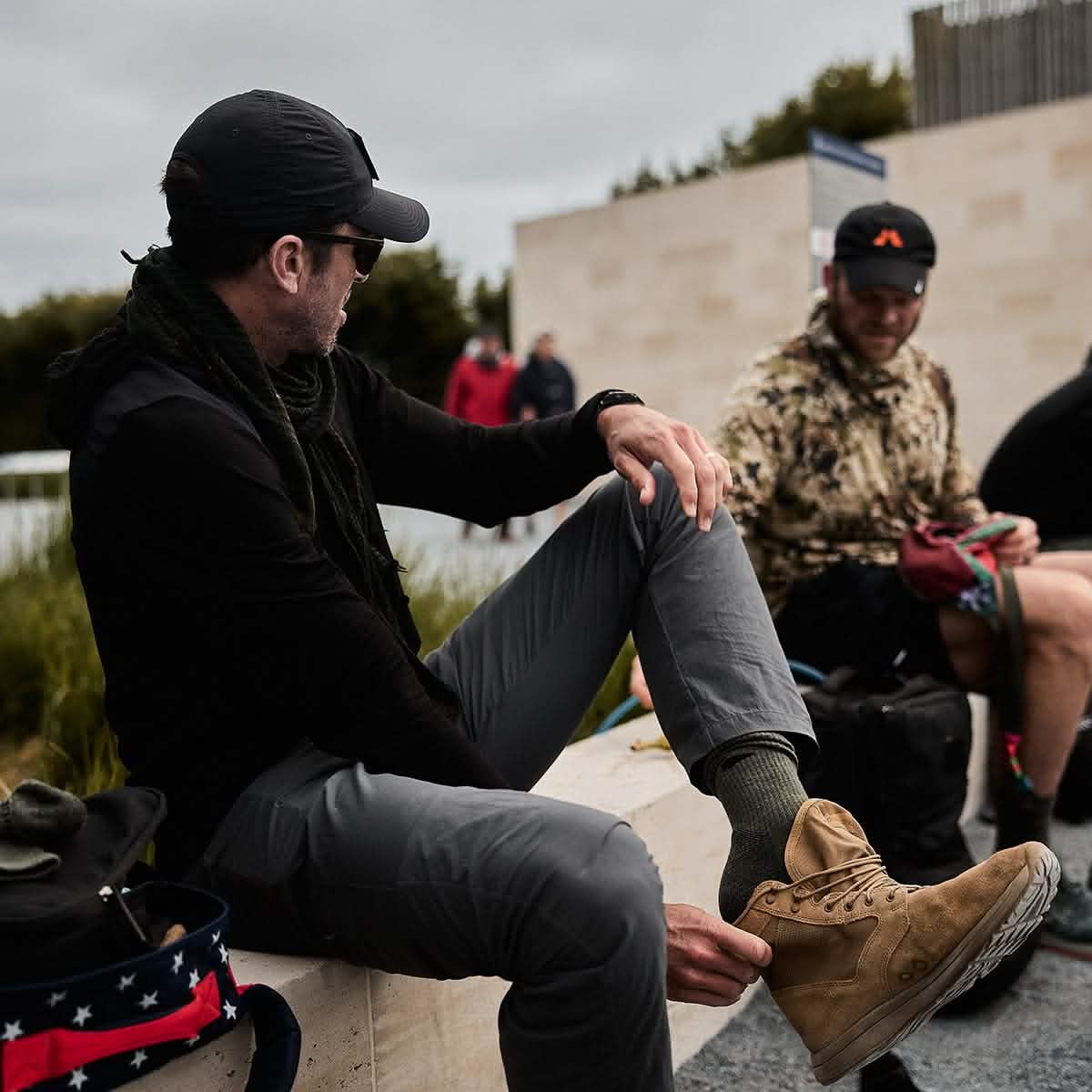 A man is seated on a wooden ledge outdoors, sporting a black cap, dark jacket, gray pants, and tan boots. His feet are snug in GORUCK's Merino Challenge Socks - Crew. He glances to his left at another person dressed in patterned clothing with a backpack nearby. The atmosphere is casual and overcast.