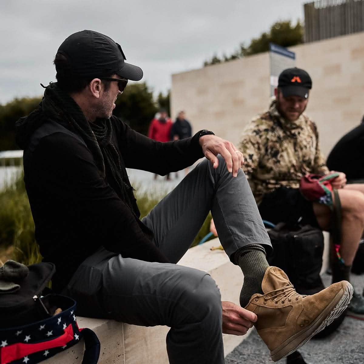 Man wearing ranger green merino wool socks, gray pants, and tan boots sitting outdoors with others in casual rucking gear