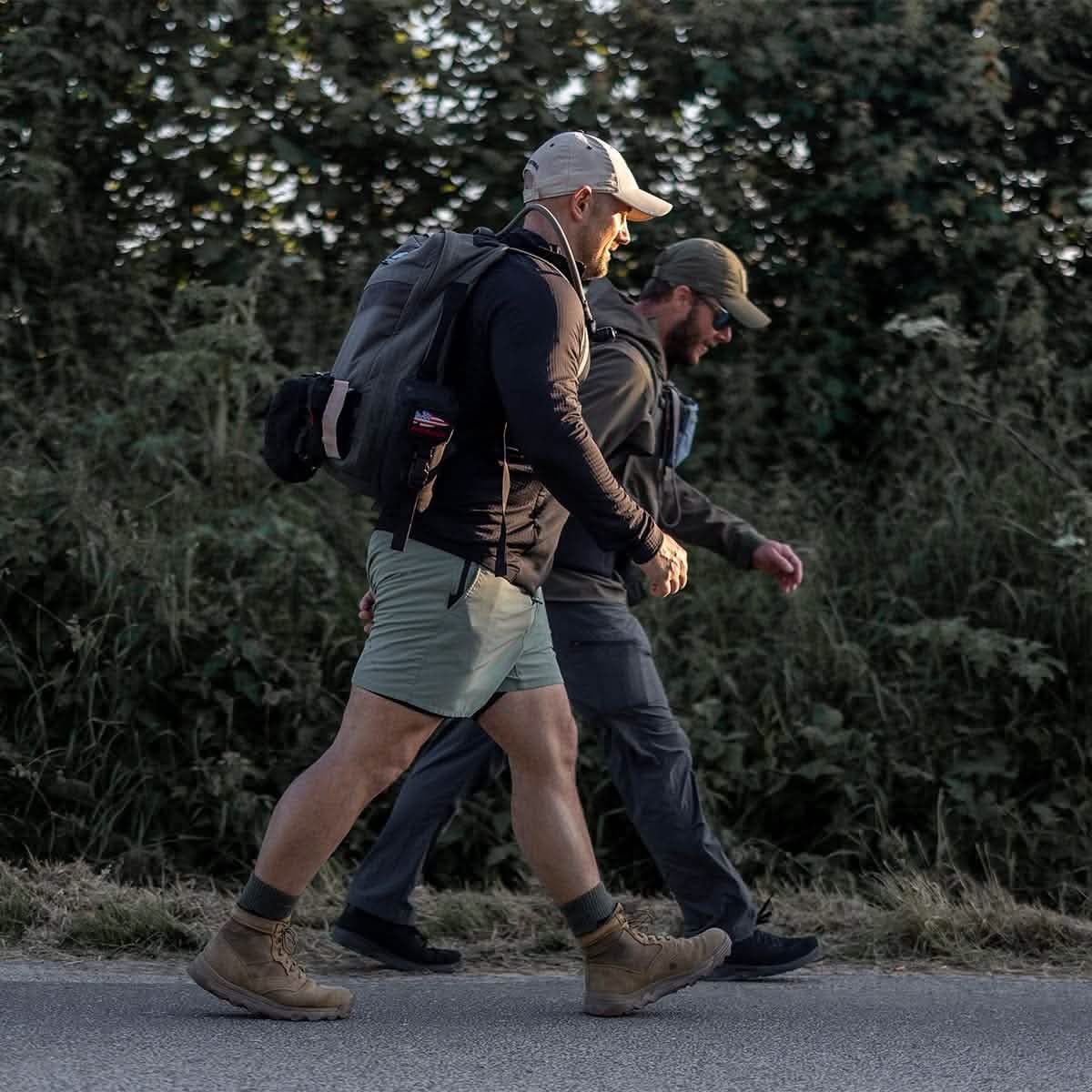 Two people walk side by side along a road, their hats and casual outdoor attire complementing the sturdy backpacks on their backs. Tall greenery lines the roadside, while the sun casts long shadows on their GORUCK Merino Challenge Socks - Crew, ensuring comfort with every step.
