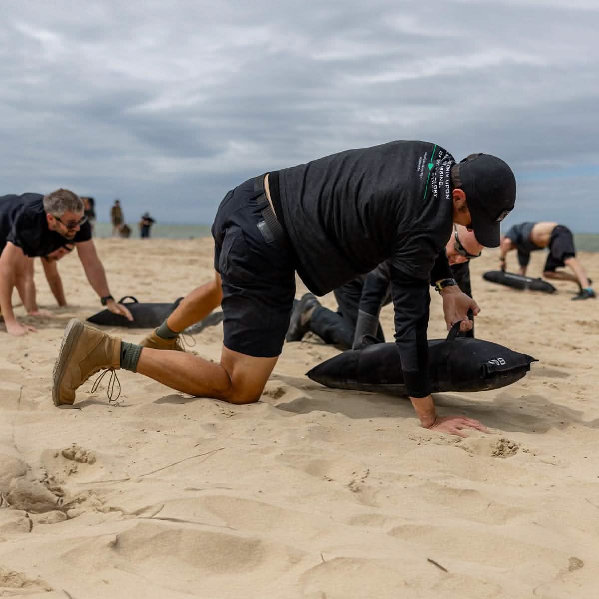 A group of people are exercising on a sandy beach, dressed in athletic clothing and Merino Challenge Socks - Crew by GORUCK, as part of a team-building activity or fitness challenge beneath a cloudy sky. In the foreground, one individual is down on all fours with a weighted bag, confidently relying on their Scars Lifetime Guarantee gear.