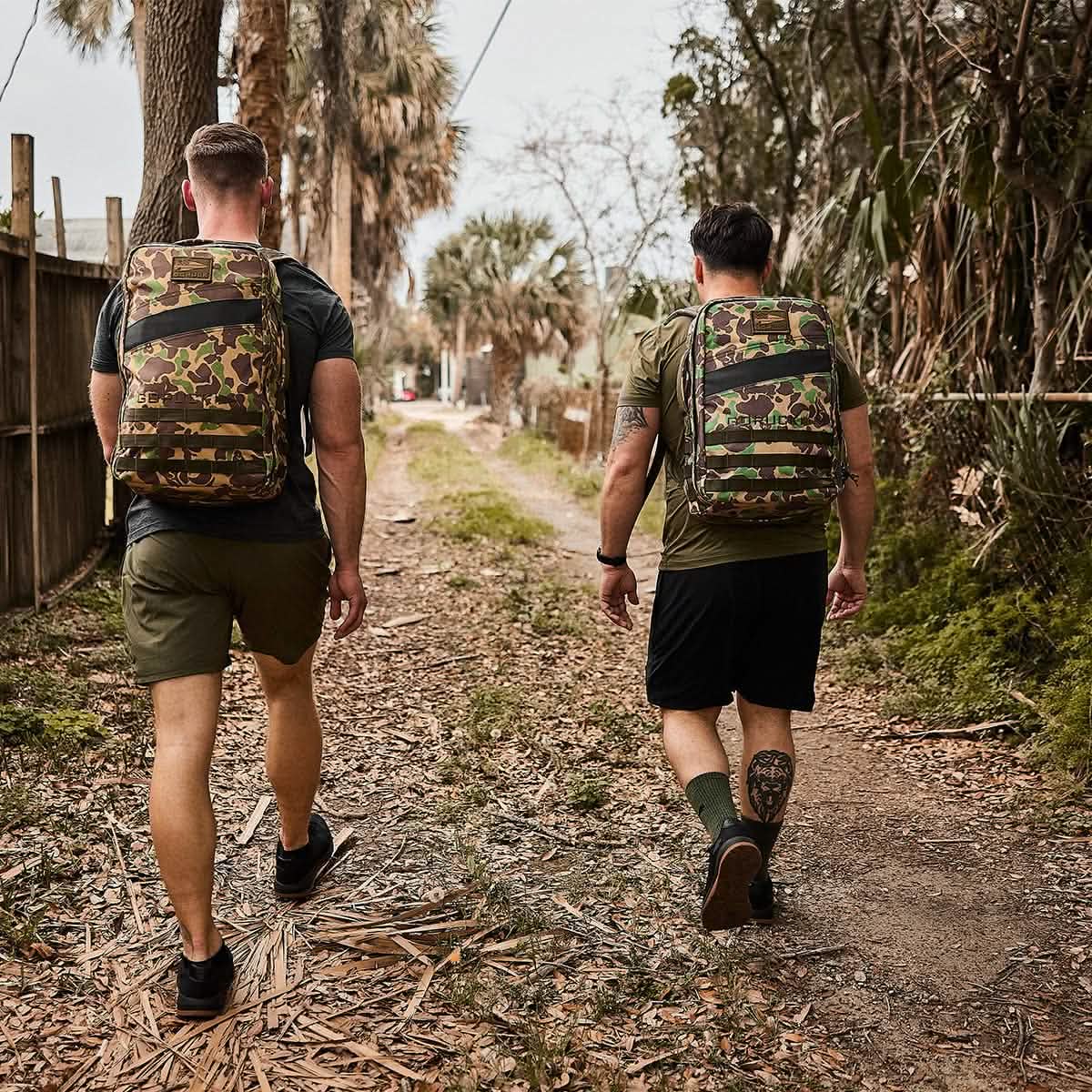 Two people walking on a dirt path surrounded by trees and a wooden fence, their attire hinting at adventure. Clad in green and black with camouflage backpacks, they tread through dry leaves, perhaps in a forest or rural area, with the comfort of GORUCK's Merino Challenge Socks - Crew cushioning each step.
