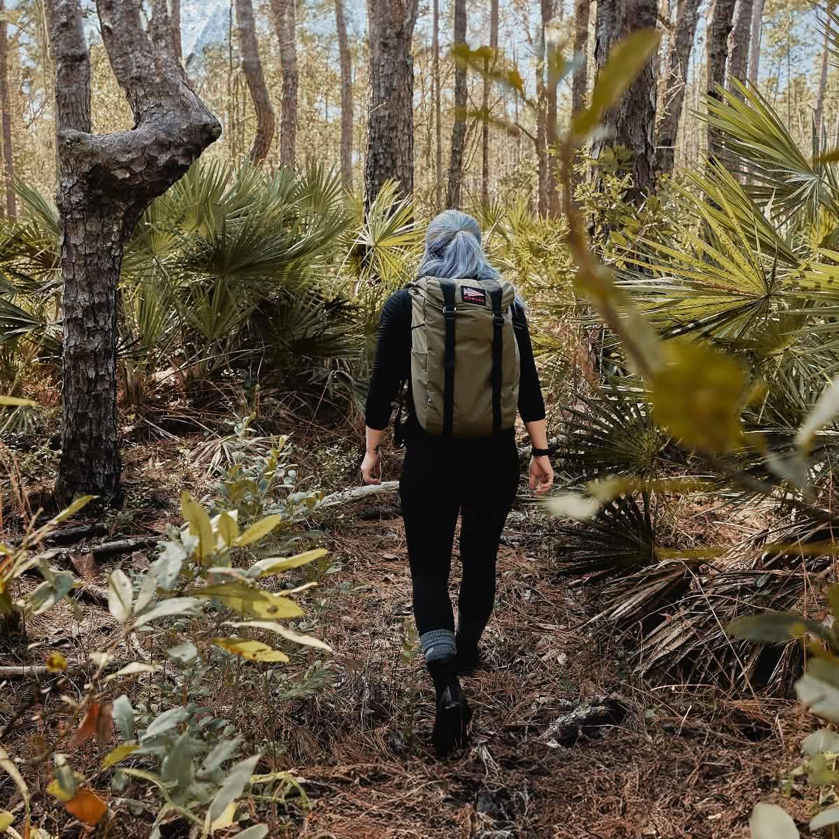 Person hiking through forest with olive green GORUCK backpack and black outfit in dense natural setting