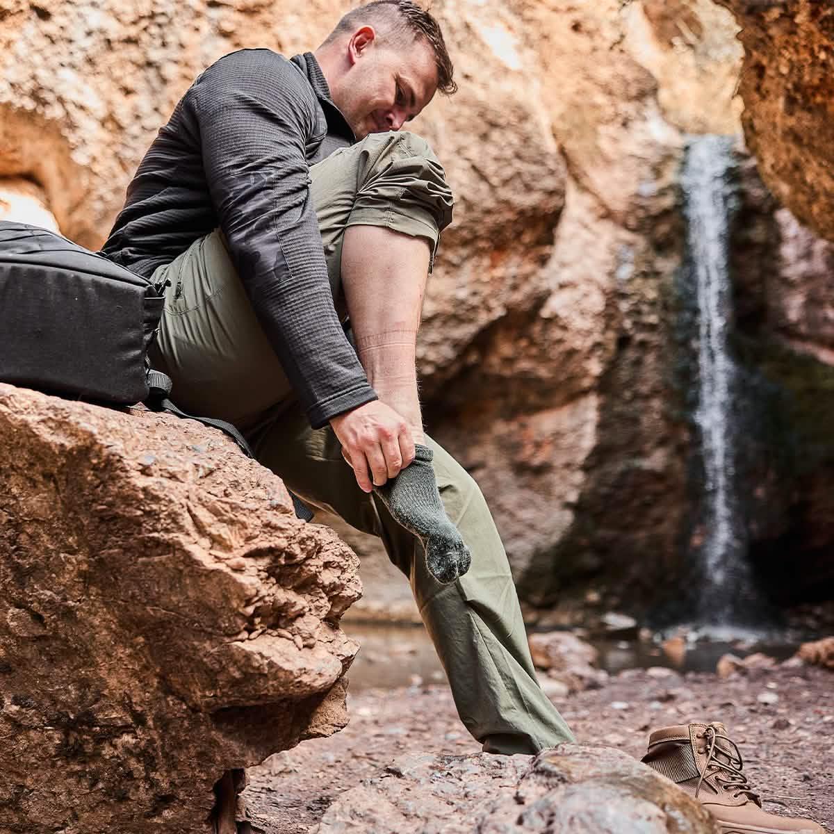 Man sitting on rock near waterfall putting on green merino wool hiking socks with hiking boots nearby