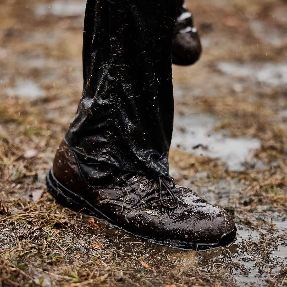 A person wearing muddy dark brown leather dress shoes and soaked black pants, paired with the Merino Challenge Socks - Crew by GORUCK, stands in a puddle on a dirt and grass surface during a rainy day.