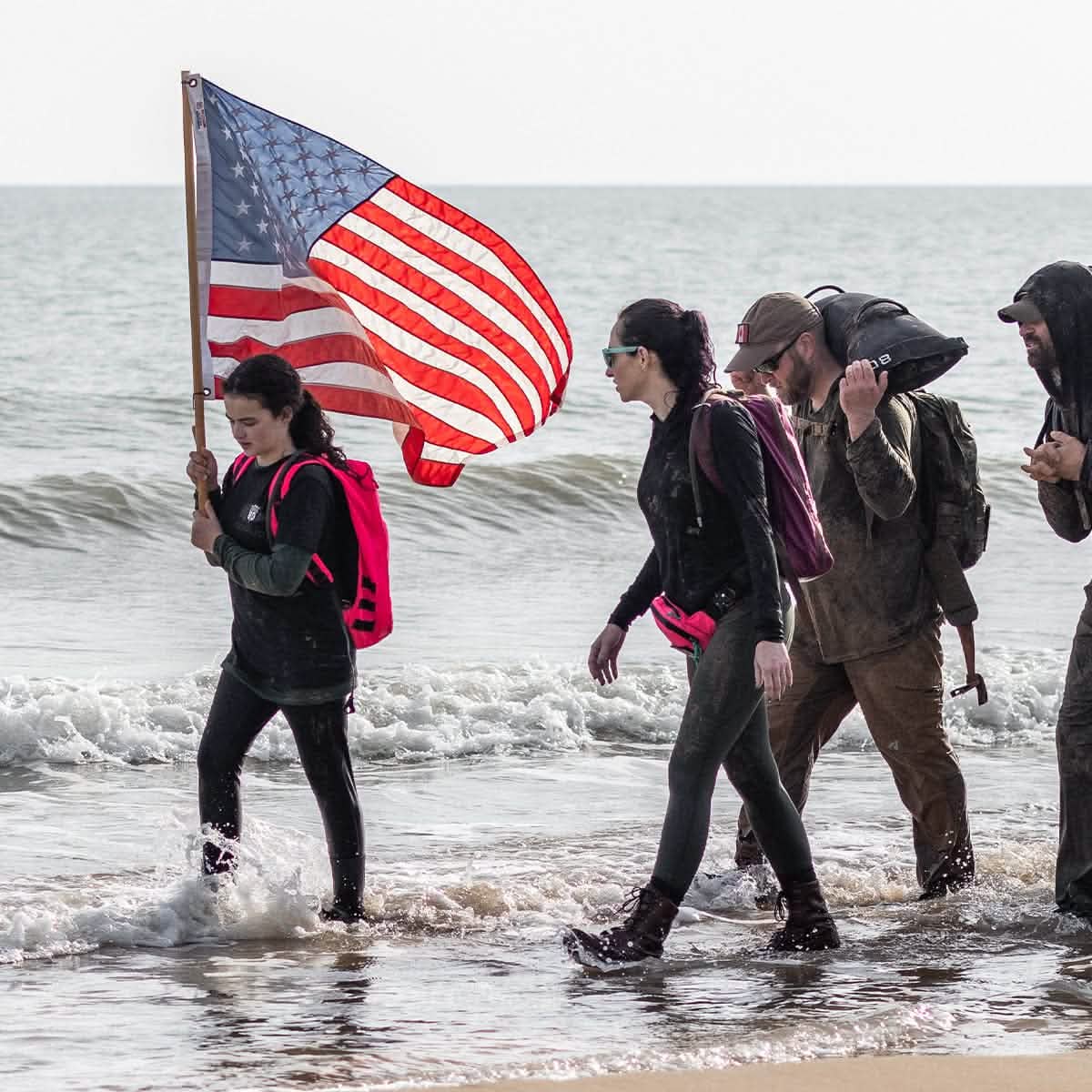 A group of people walking through the surf on a beach, with one person carrying a large American flag made in the USA. Dressed in casual outdoor clothing, another individual wears GORUCK's Merino Challenge Socks - Crew and carries a heavy-looking bag over their shoulder as ocean waves gently lap against them.