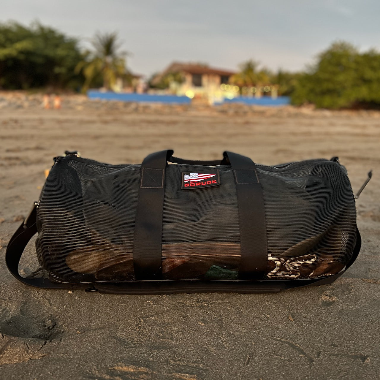 The Mesh Duffel Bag in black is ideal for beach outings, shown resting on sandy shores with blurred trees and a house in the background.