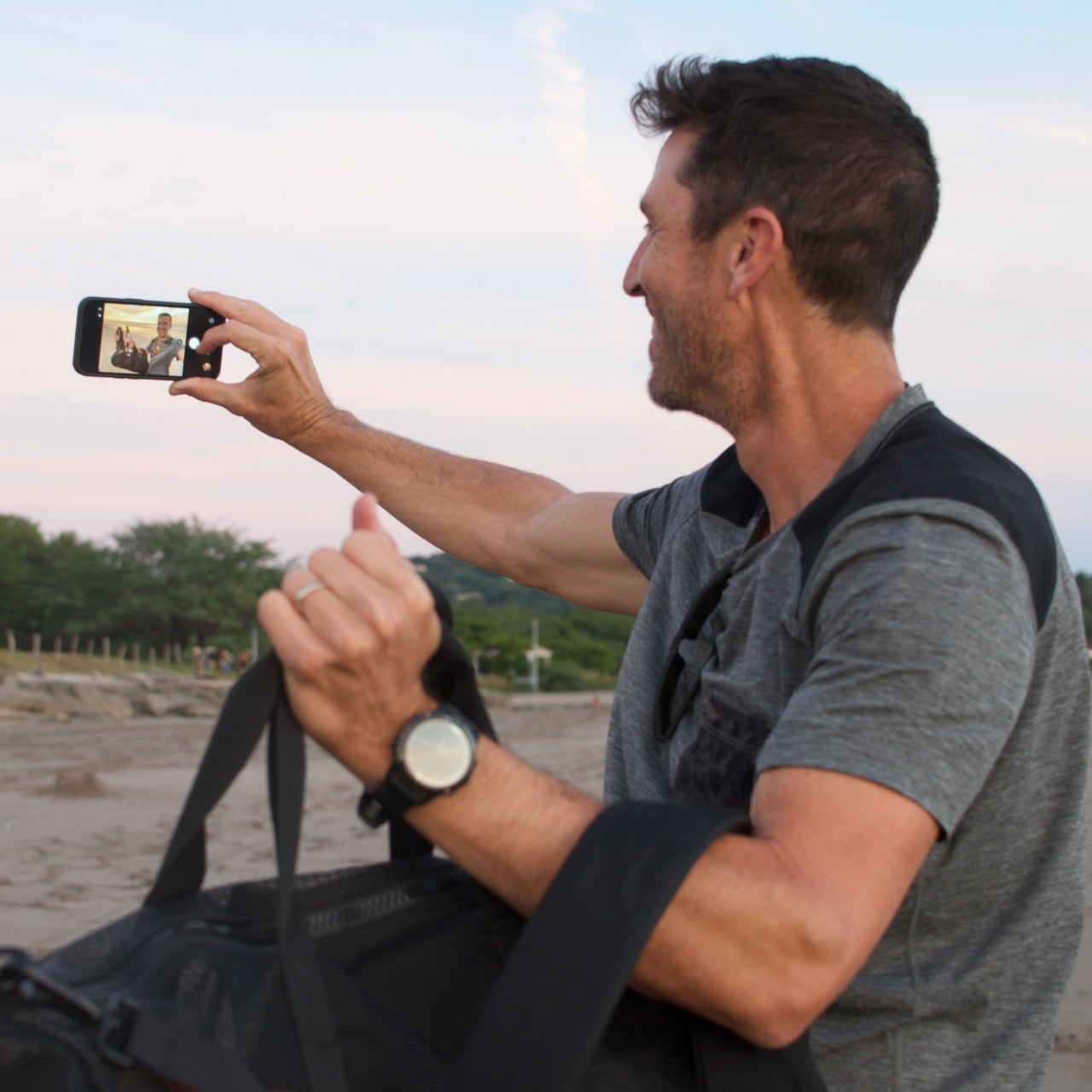 A man smiles and takes a selfie outdoors, carrying the Mesh Duffel Bag over his shoulder—ideal for storing wet clothes after an active day out.