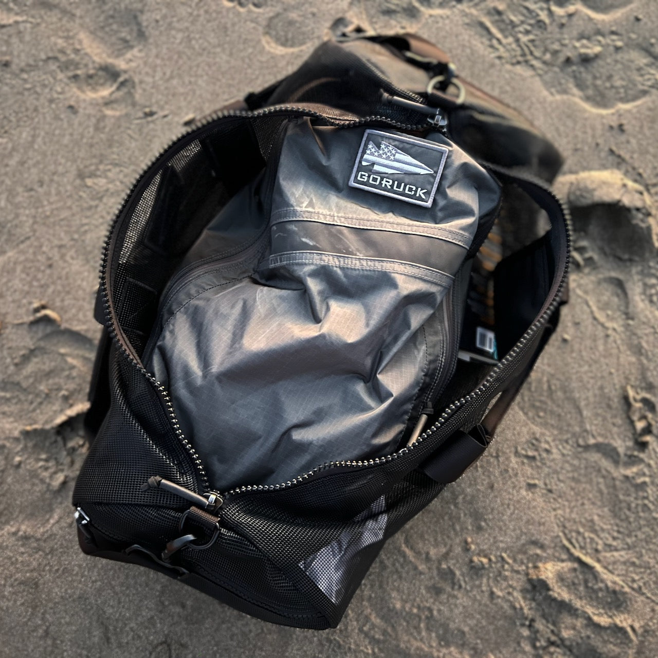 An open black Mesh Duffel Bag sits on sand with a packed jacket inside, displaying the GORUCK logo.