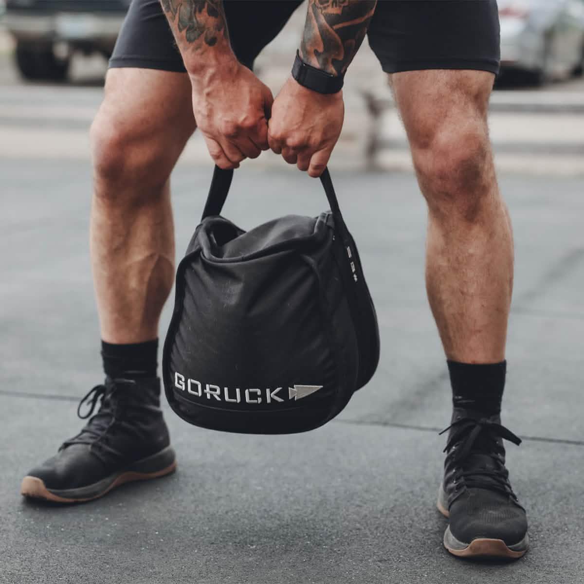 Person holding a black GORUCK weighted sandbag during outdoor fitness training, wearing black shorts and black shoes