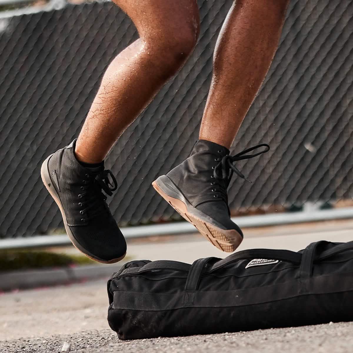 Person jumping in black GORUCK boots over a black rucking gear bag on pavement with chain-link fence background