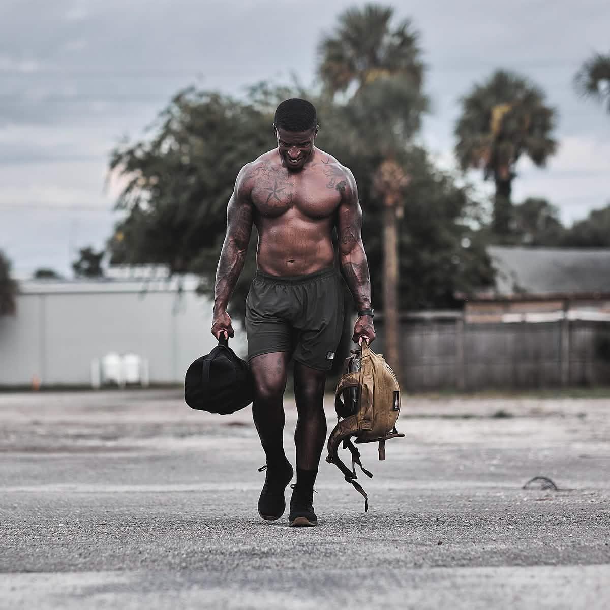 Muscular shirtless man walking outdoors carrying GORUCK rucking bags with palm trees in background