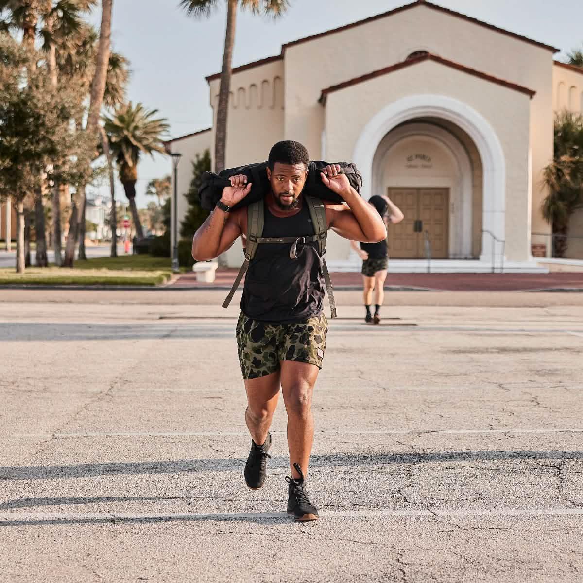 Athletic man wearing camo shorts and black tank top carrying a weighted rucking bag outdoors near palm trees and a beige building