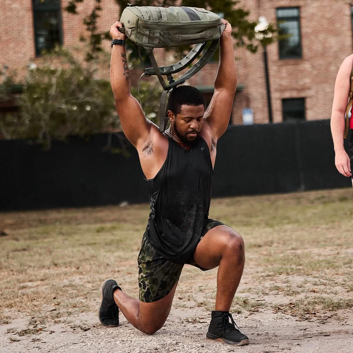 Man in black tank top and camouflage shorts lunging outdoors while holding GORUCK bag overhead during workout