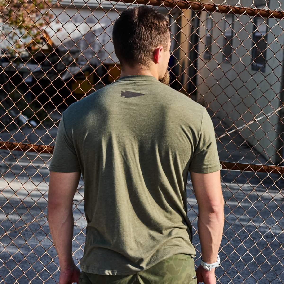 Man wearing military green GORUCK t-shirt and black frogskin camo shorts standing by a rusted chain link fence