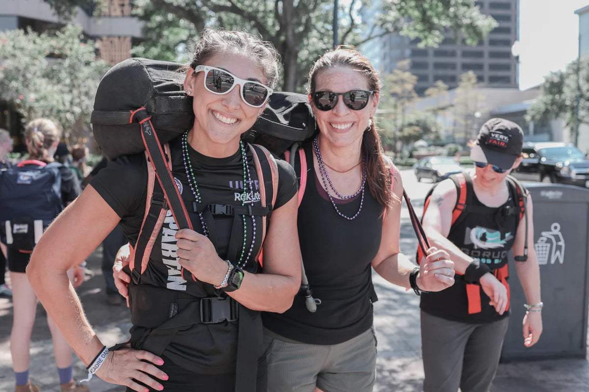 Two smiling women in athletic wear pose outdoors with backpacks, sunglasses, and race bibs at the inclusive MOTHERUCKER 5K fitness event.