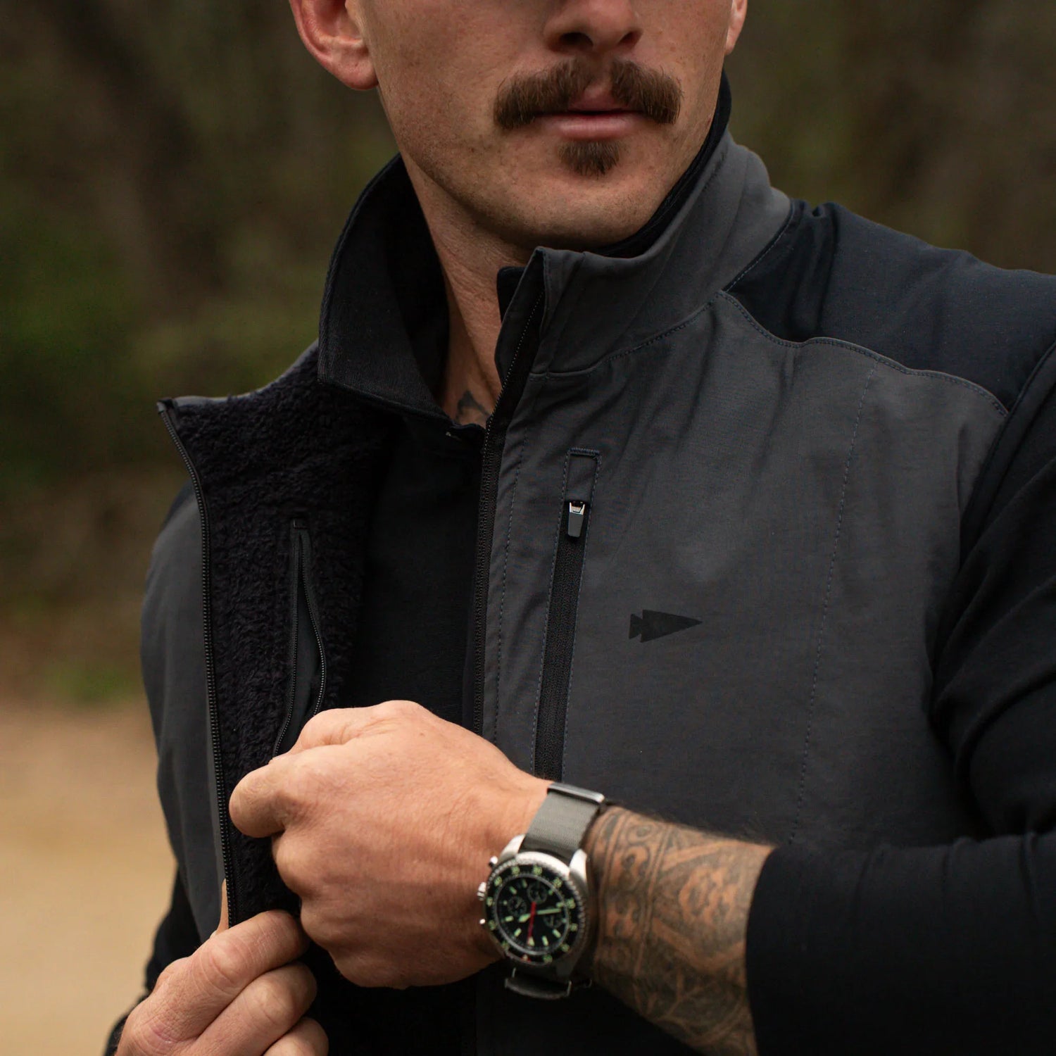 Man wearing dark GORUCK jacket with zipper and logo, sporting tactical watch and tattooed arm outdoors