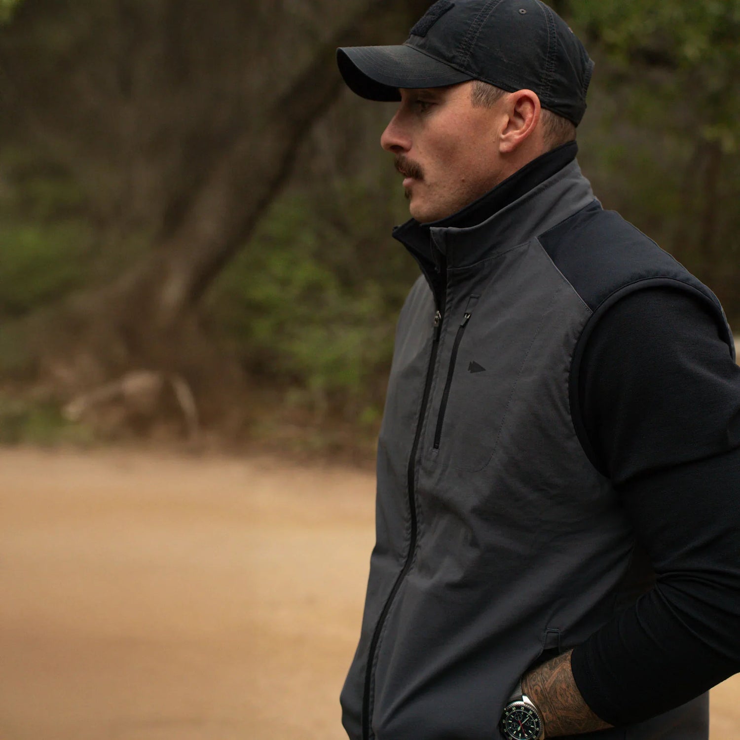 Man in black cap and rugged dark outdoor vest standing on dirt trail with wooded background