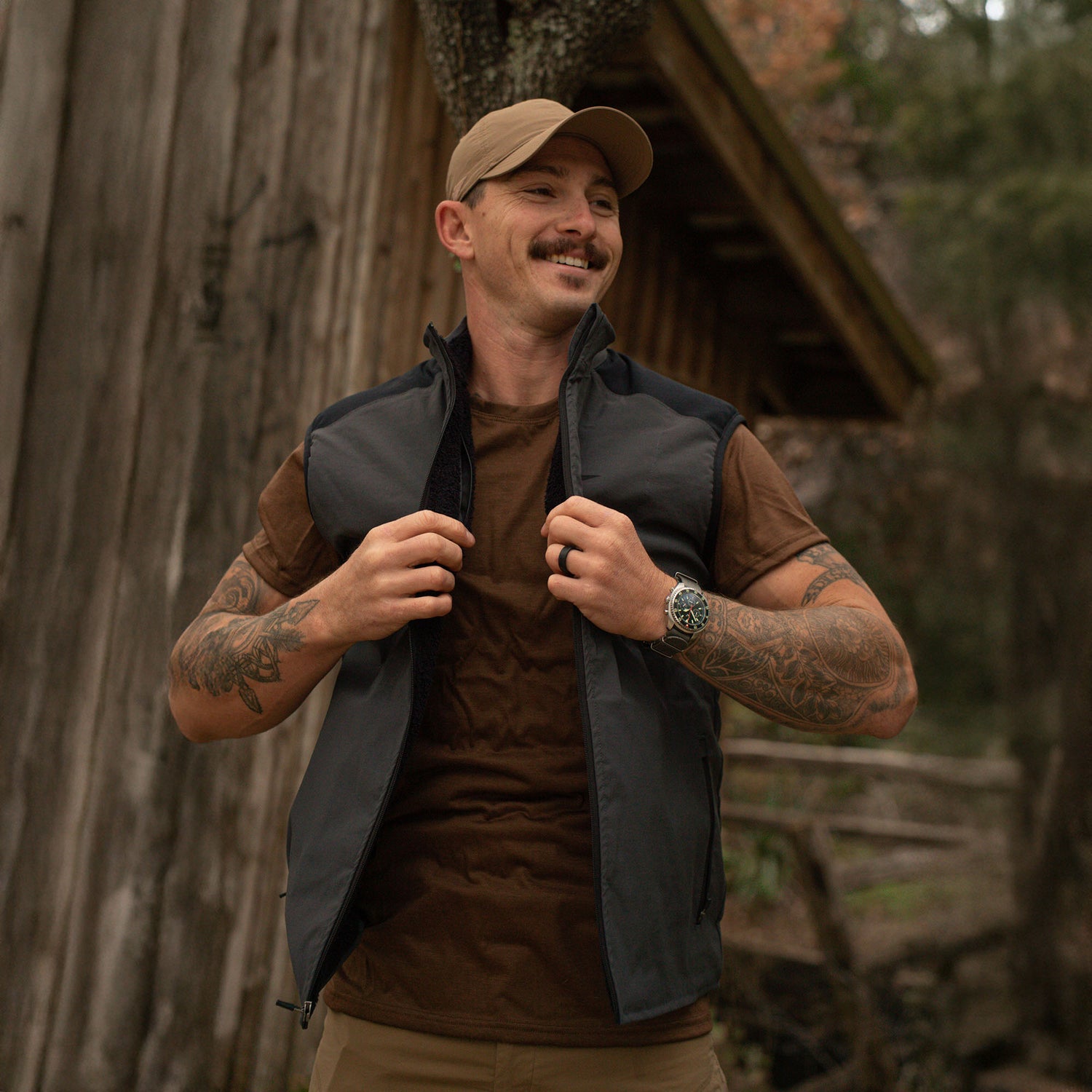 A smiling man in a cap and the Men's Vest of Power - ToughDry® + Fleece, made in the USA, stands outside by a rustic wooden building, adjusting his vest.