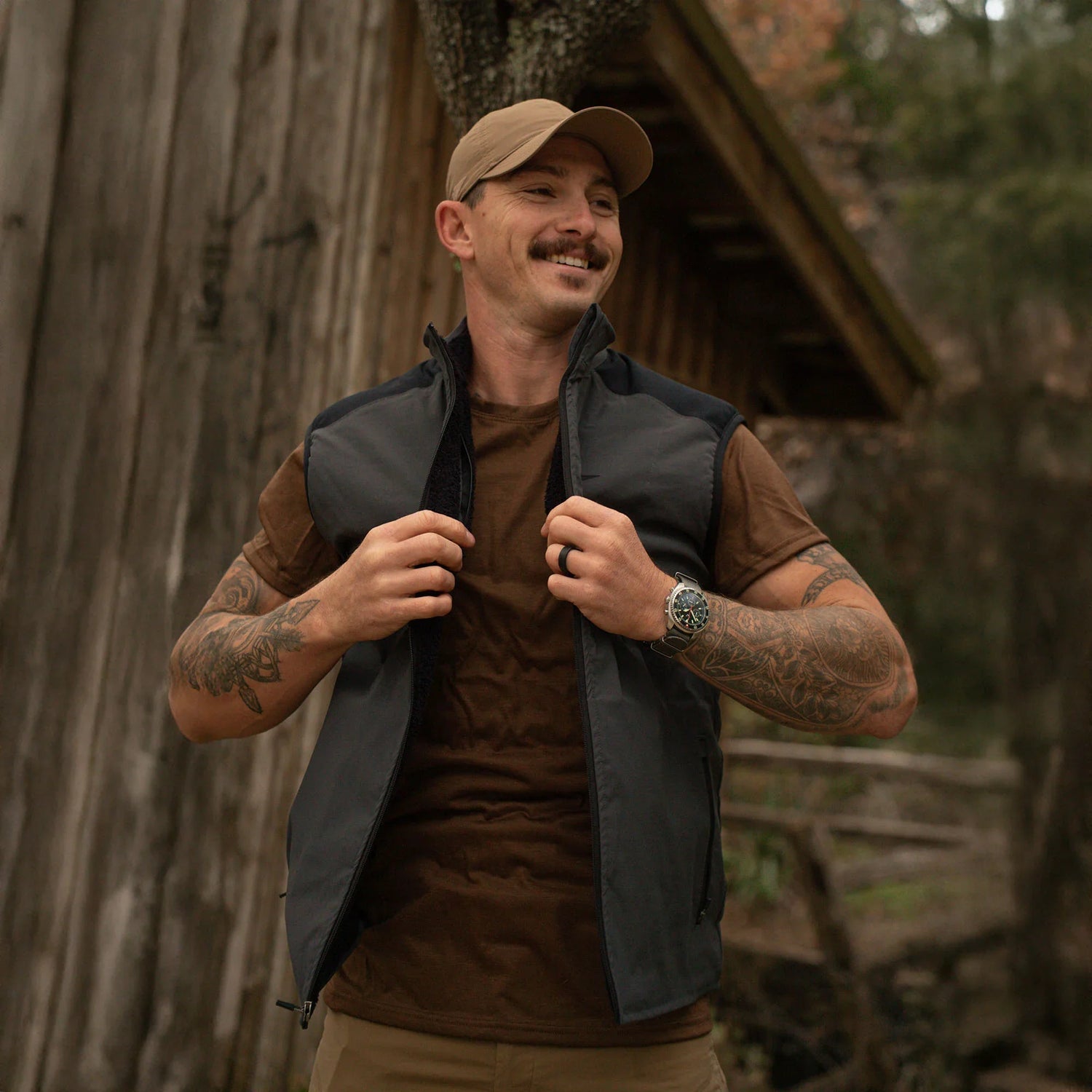 Man wearing brown cap and shirt with black tactical vest, standing outside rustic wooden cabin in forest
