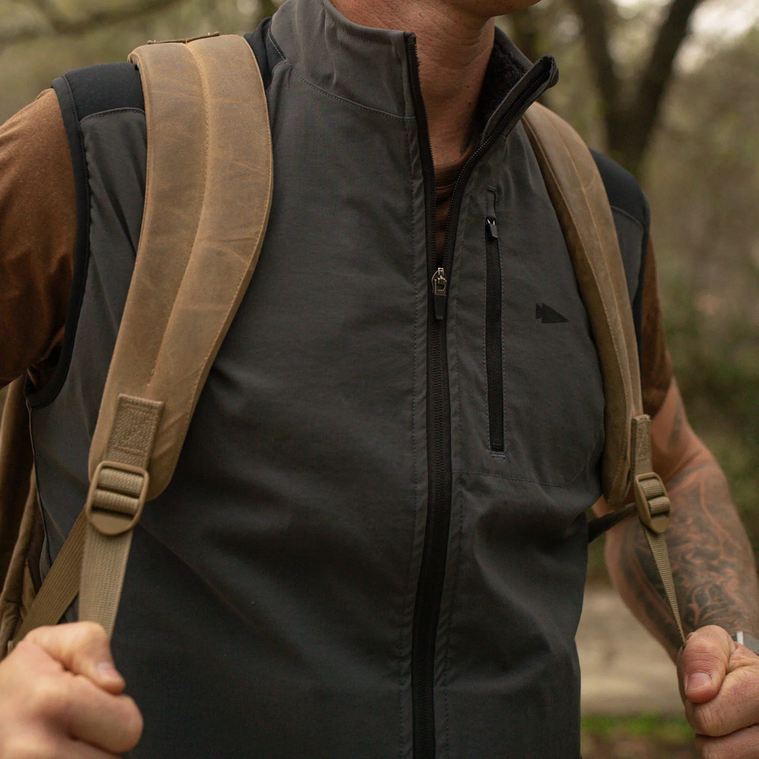 Close-up of a person wearing a black zip-up vest and tan backpack straps with outdoor background