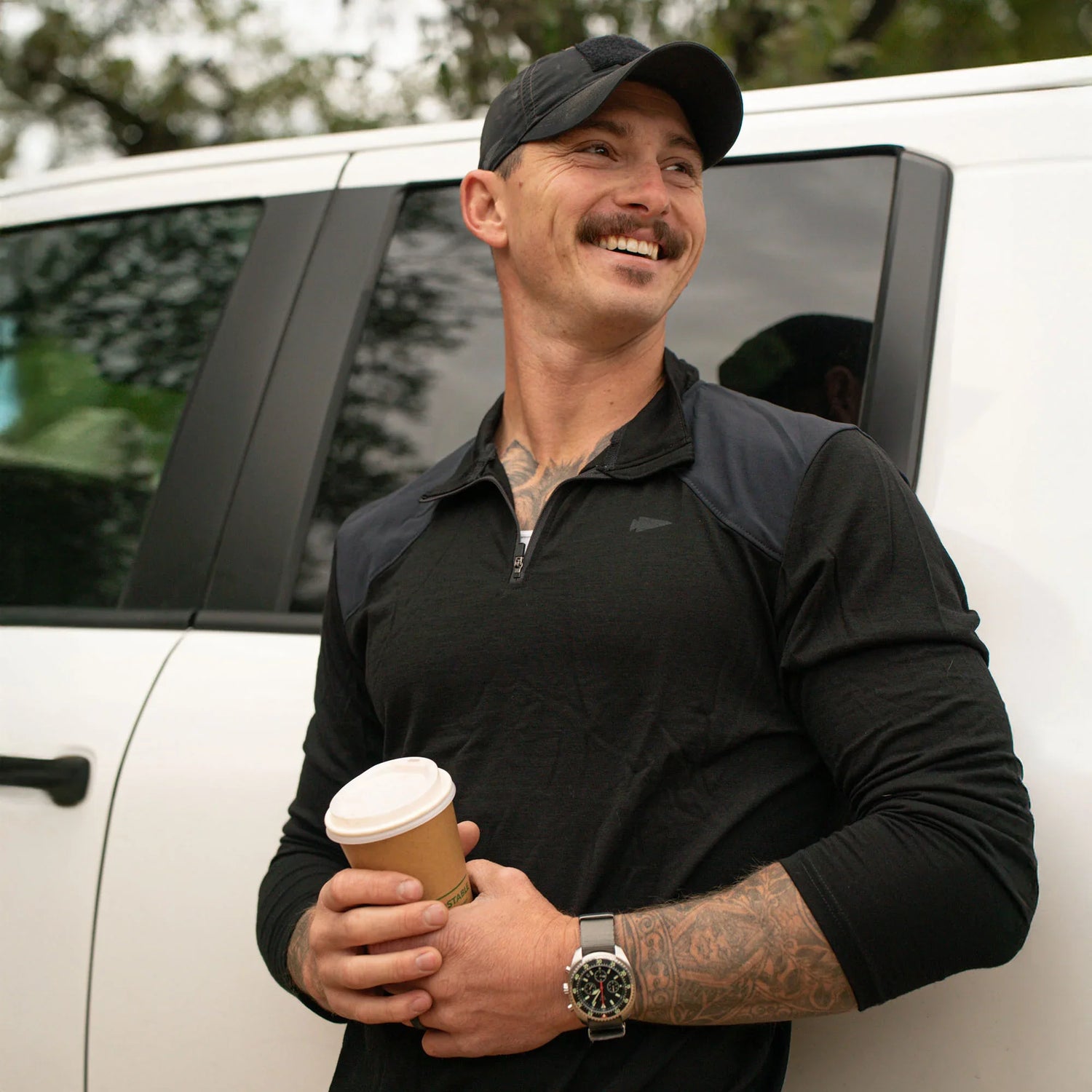 Smiling man in black GORUCK gear holding coffee, standing by a white vehicle outdoors.
