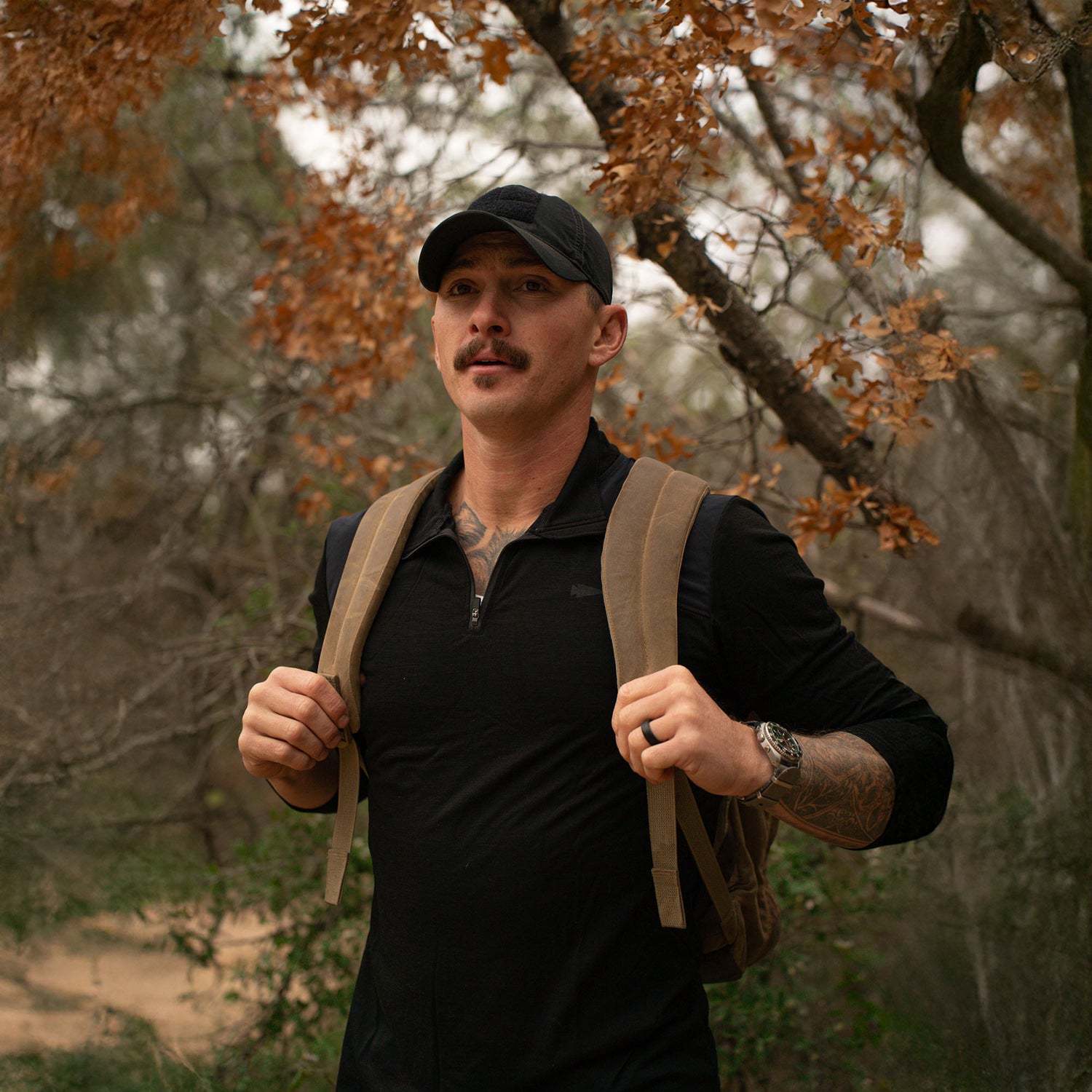 A man with a mustache and tattoos, wearing a black cap and backpack, stands outdoors among autumn trees, staying comfortable in the Men's Commando Quarter Zip - Merino Wool.