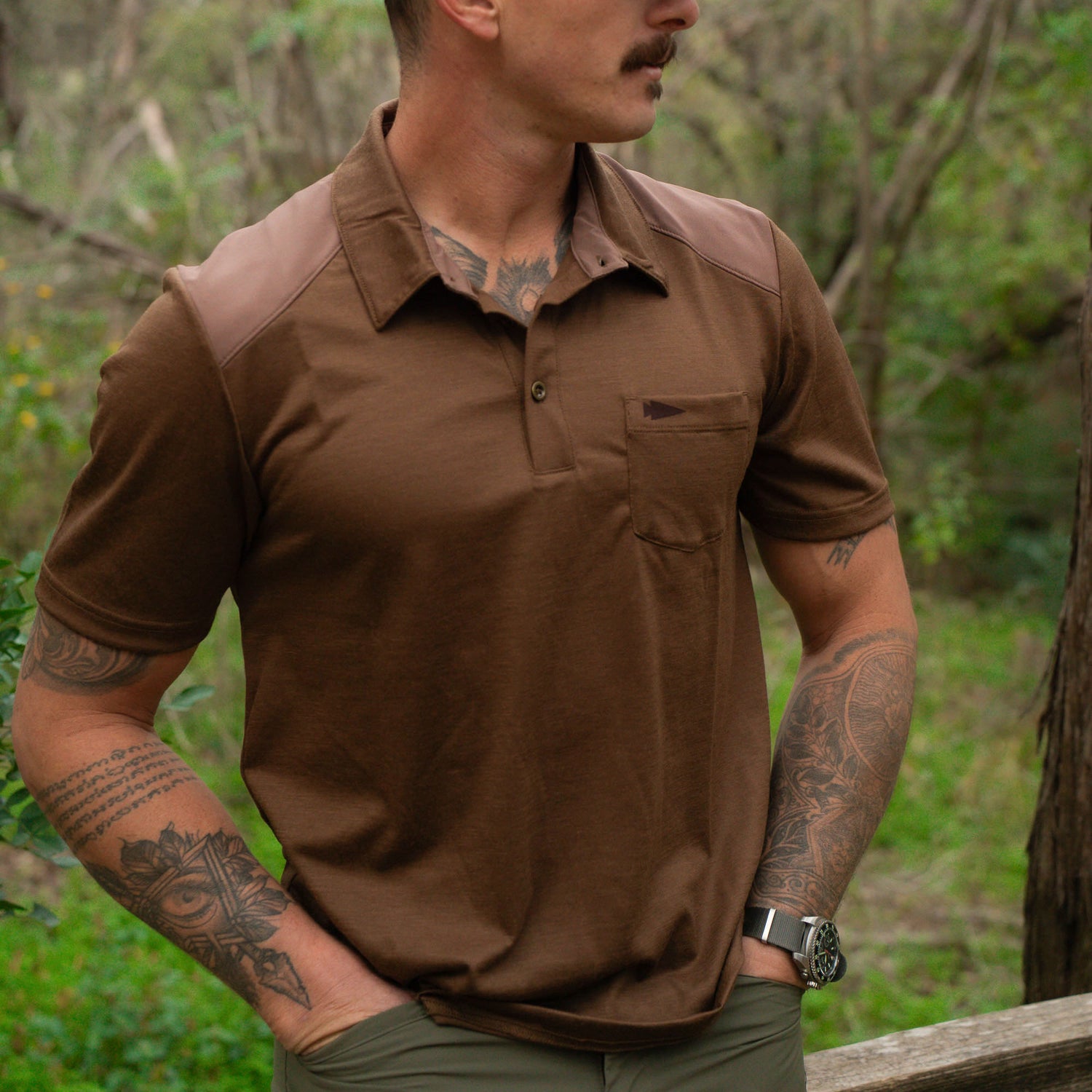 A tattooed man stands outdoors in a wooded area, wearing the Men's Commando Polo - Merino Wool in brown, with one hand resting in his pocket.