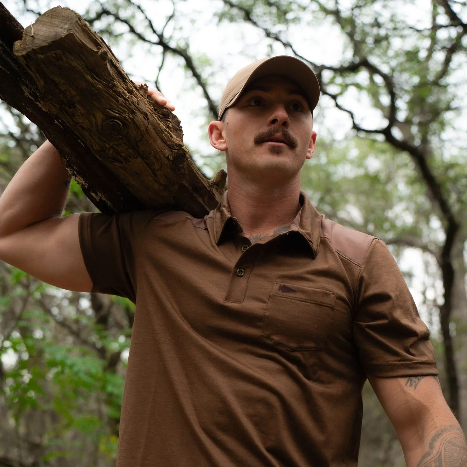 Man wearing GORUCK brown shirt and cap carrying a log in a wooded outdoor setting