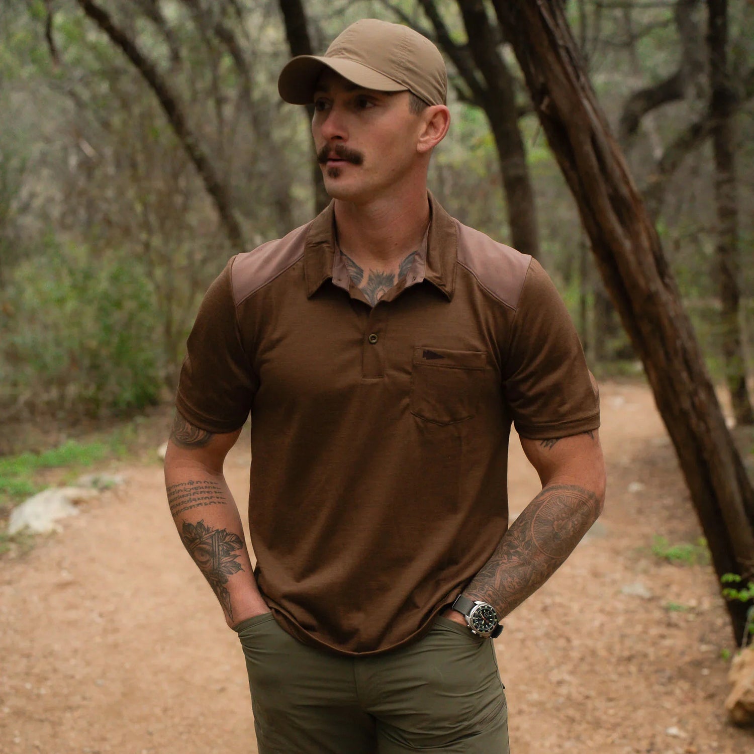 Man in brown GORUCK shirt and tan cap standing on a forest trail, showcasing rugged outdoor gear.
