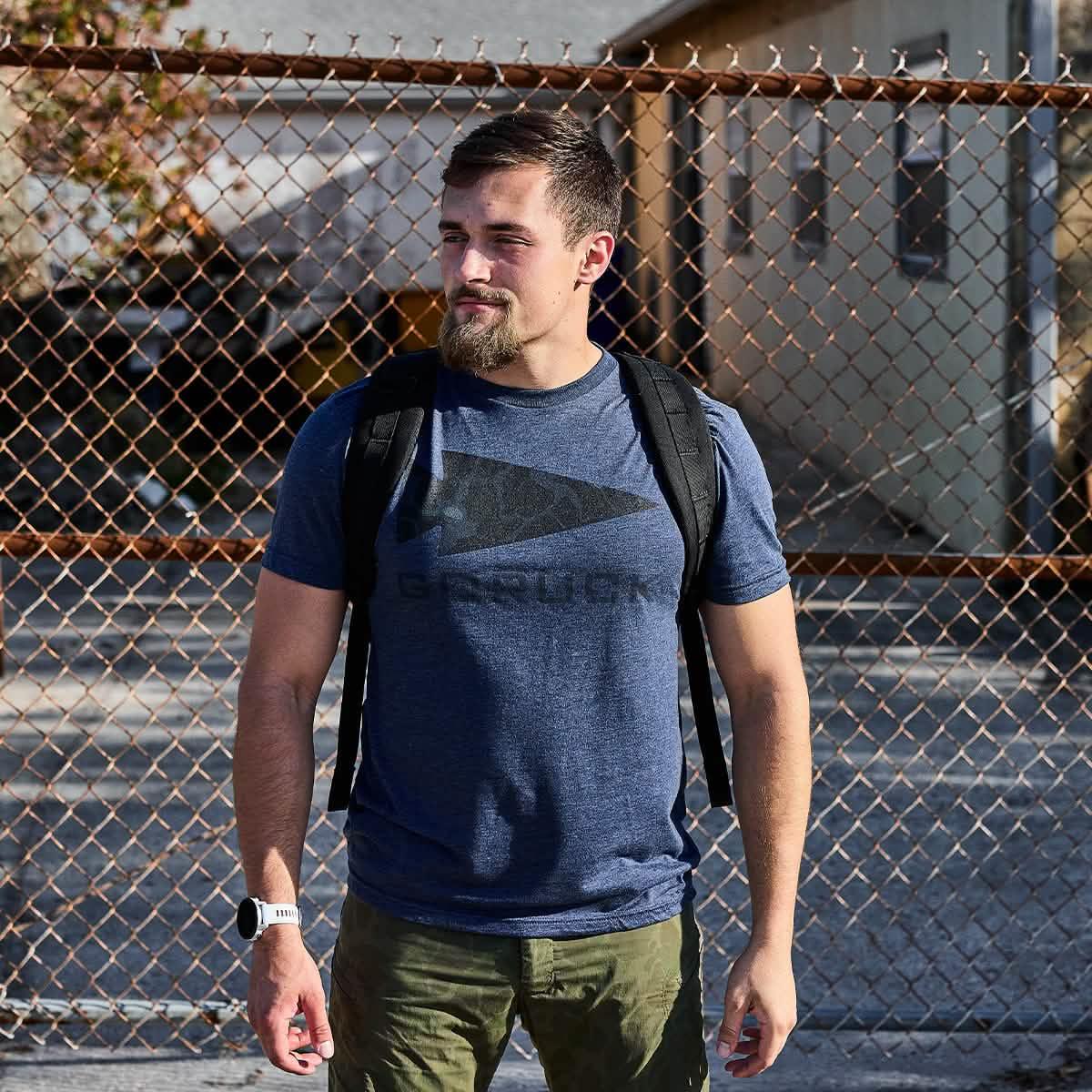 Man wearing GORUCK navy camo t-shirt and rucksack standing by a chain-link fence outdoors