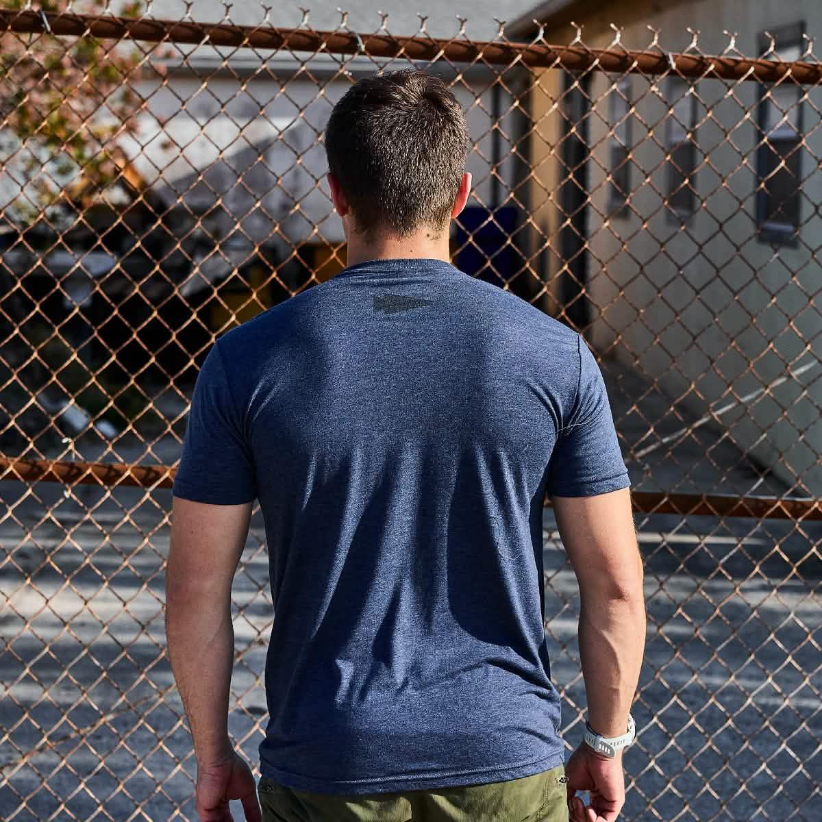 Man wearing navy blue GORUCK t-shirt standing in front of rusted chain-link fence outdoors