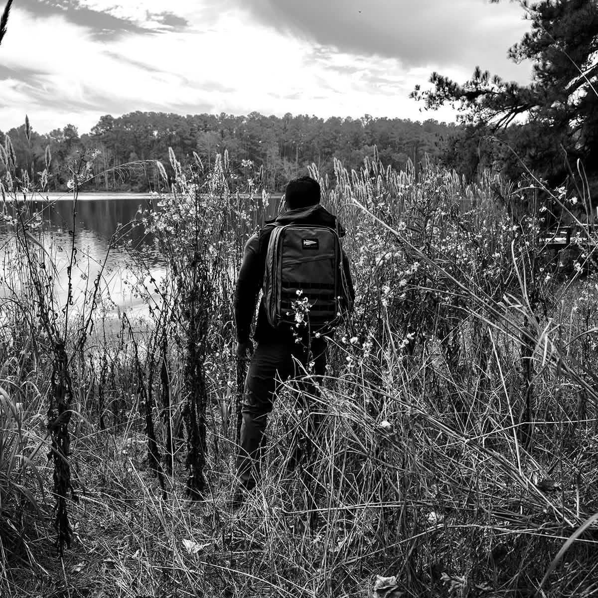 Person wearing GORUCK Black Dyneema 34L backpack standing amid tall grass by a lake in nature