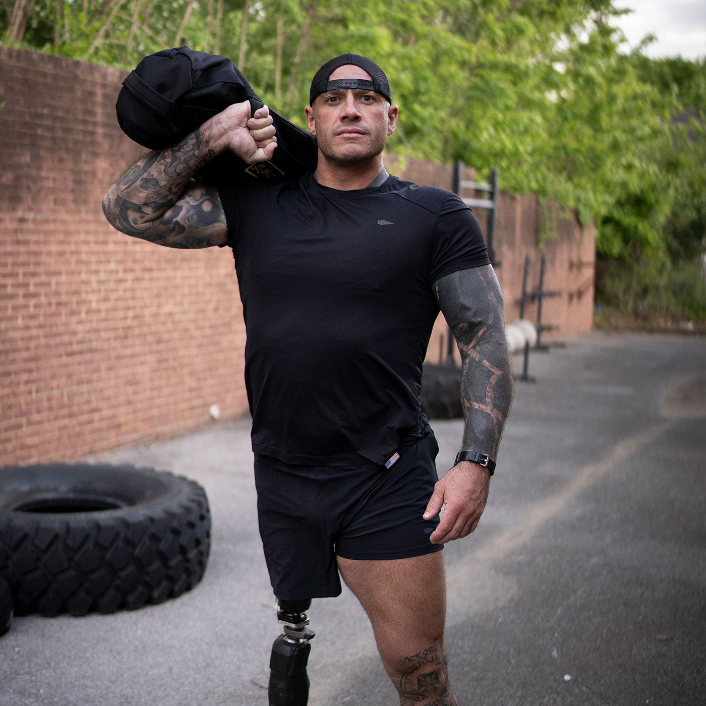 A muscular man with a prosthetic leg carries a sandbag over his shoulder outdoors near tires and gym equipment.