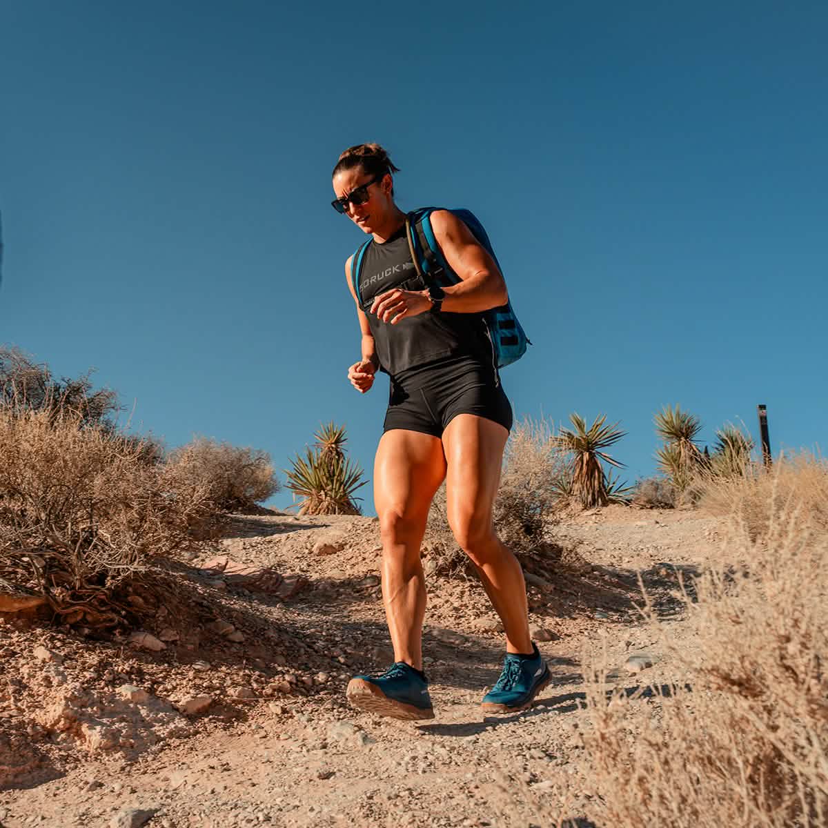 Under a clear blue sky, a person in athletic gear and sunglasses hikes a rugged trail, enjoying the comfort of GORUCK's Merino Challenge Socks - Ankle. Carrying a backpack, they navigate the path surrounded by dry shrubs and desert plants.