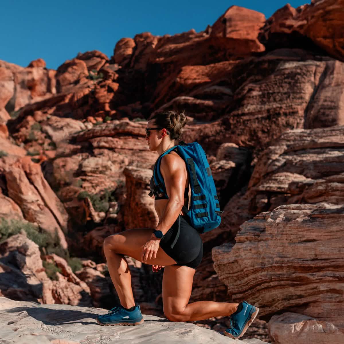 Clad in athletic attire and carrying a blue backpack, the individual wearing GORUCK's Merino Challenge Socks - Ankle pauses mid-lunge on rugged terrain. Set against a breathtaking backdrop of red rock formations beneath a clear blue sky, they embody the adventurous spirit of those who appreciate quality gear crafted in the USA.