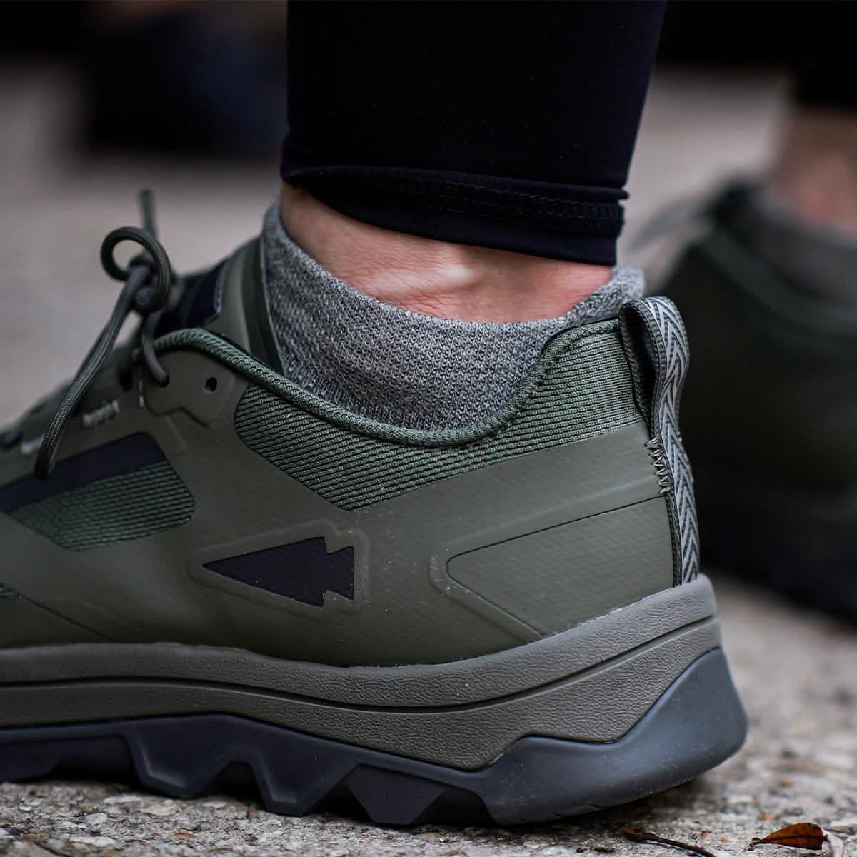 Close-up of a person wearing Merino Challenge Socks - Ankle by GORUCK alongside green hiking shoes with grey accents. The individual stands on a textured outdoor surface, displaying the shoe's thick sole and rugged design, indicating its suitability for outdoor activities and assurance through the Scars Lifetime Guarantee.