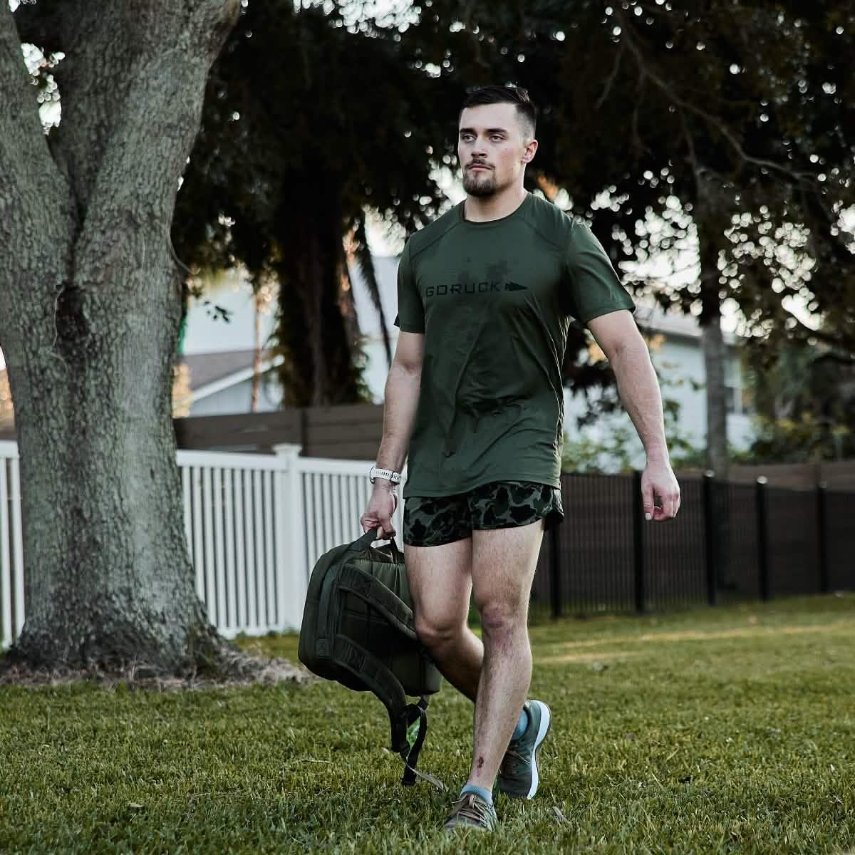 Man wearing olive green GORUCK t-shirt and camo shorts walking on grass carrying a backpack outdoors