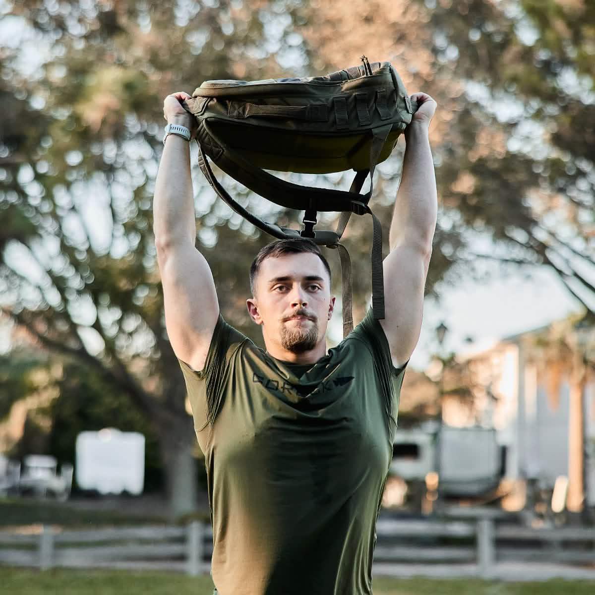 Man outdoors wearing olive GORUCK tough mesh tee lifting a green rucksack above his head in a park setting