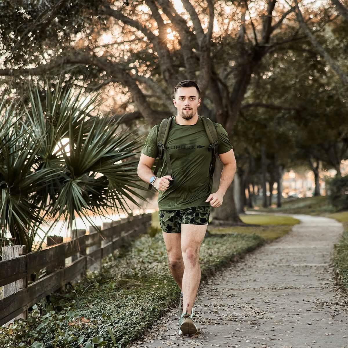 Man jogging on leafy park path wearing olive GORUCK tough mesh tee, camo shorts, and a backpack at sunset