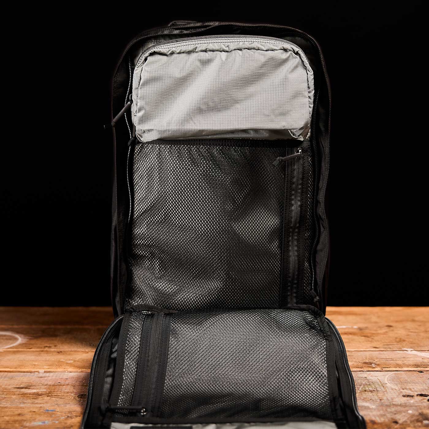 Open black backpack with mesh pockets, lying flat on a wooden surface against a black background.